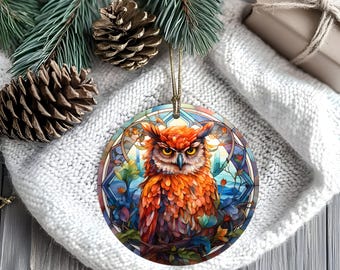 May include: Round Christmas ornament featuring a detailed illustration of an owl with orange and brown feathers, set against a stained-glass style background with blue and green hues. The ornament has a gold string for hanging.