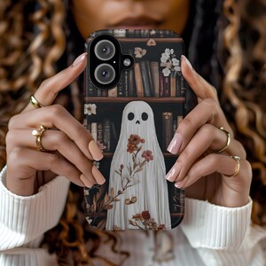 Pode incluir: Um fantasma branco com um design floral está em pé na frente de uma estante cheia de livros. O fantasma tem um rosto de caveira e está rodeado de flores. A estante é marrom escura e tem um fundo branco.