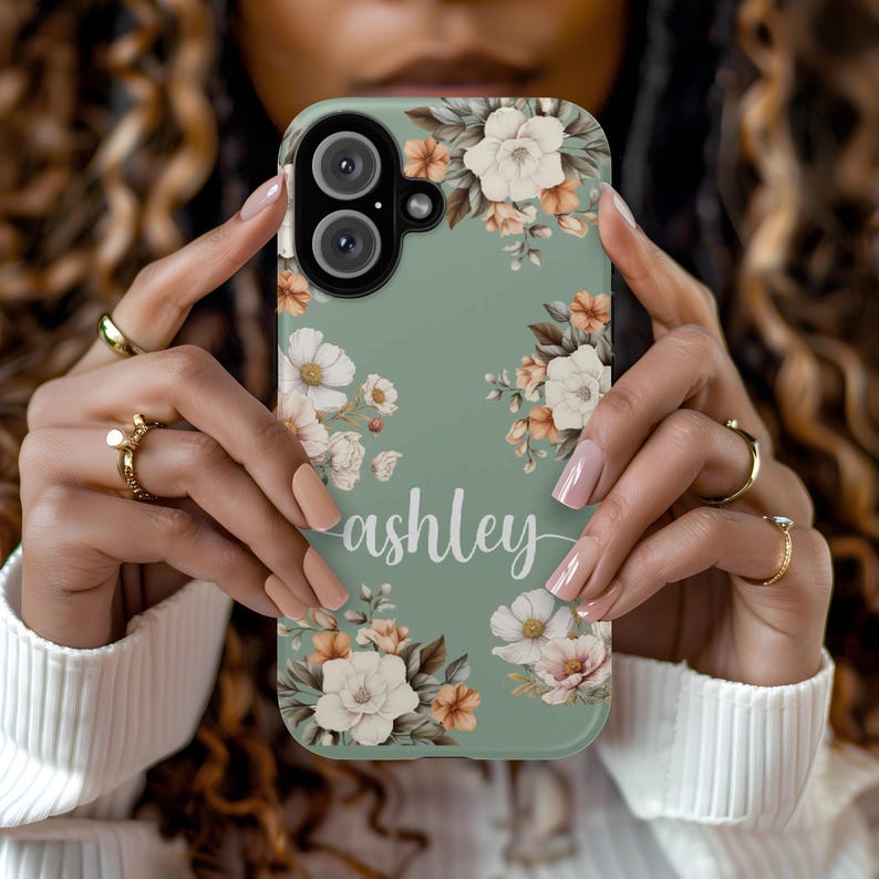 May include: A sage green phone case with a floral design and the name "ashley" printed on it.