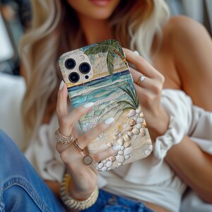 May include: A phone case featuring a beach scene with a palm tree, ocean waves, and seashells. The case has a mosaic-like design with shades of blue, beige, and green. The phone case is held by a person.
