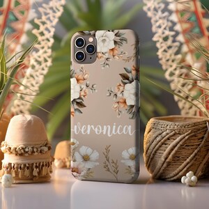 May include: A beige phone case with a floral design and the name "Veronica" printed on the bottom. The flowers are white, pink, and orange.