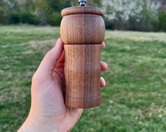 May include: A wooden pepper grinder with a dark brown finish. The grinder has a simple design and is held in a hand against a green grassy background.