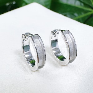 May include: A pair of silver-toned hoop earrings featuring a polished outer edge and a textured, matte-finish center band. These circular earrings have a hinged closure mechanism, providing a secure fit while maintaining a clean, metallic appearance.