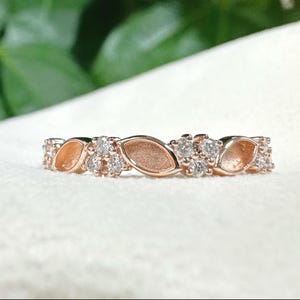 Op de afbeelding: Roségouden ring met een herhalend patroon van marquise-vormige elementen en clusters van kleine, ronde, heldere edelstenen. De ring is geplaatst tegen een zachte witte achtergrond, met een vage groene achtergrond.