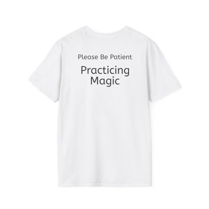 May include: White t-shirt with the text "Please Be Patient Practicing Magic" in black. The shirt has short sleeves and a crew neck. The text is centered on the front of the shirt.