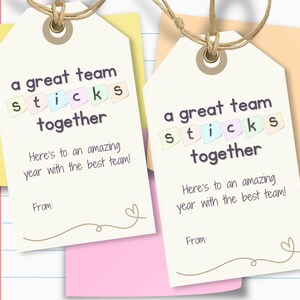 May include: Two gift tags with the text "a great team sticks together." The tags are cream-colored with a brown string and a heart design. The tags are on top of yellow and pink sticky notes. The text "Here's to an amazing year with the best team!" is also visible.