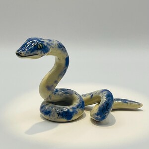 May include: A ceramic snake figurine in a coiled pose. The snake has a cream-coloured body with blue speckled patterns. The head is raised, and the eyes are visible. The figurine is on a white surface.