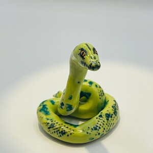 May include: A coiled ceramic snake figurine. The snake is primarily yellow-green with darker green spots and markings. The head has orange eyes and a detailed, textured appearance. The figurine is on a white surface.