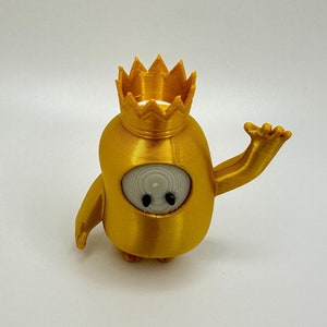 May include: A gold-colored 3D printed figurine of a character wearing a crown. The character has a white face with black eyes and a small mouth. The character is waving with its right arm.