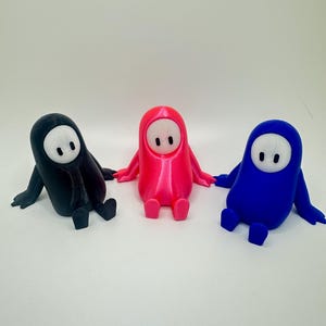 May include: Three 3D printed figurines of characters from the game Fall Guys. The figurines are sitting down with their arms outstretched. The figurines are black, pink, and blue.
