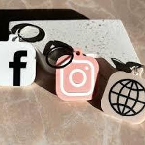 以下が含まれることがあります： Facebook、Instagram、地球儀のアイコンが描かれた正方形のチャームが付いたキーホルダーが3つ。チャームは白、ピンク、ベージュで、黒いアウトラインが描かれている。