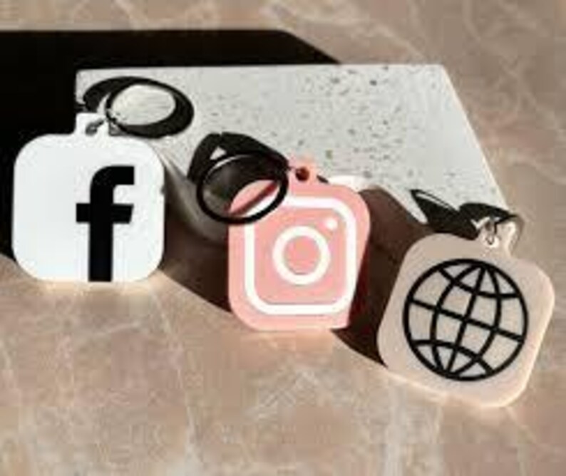 以下が含まれることがあります： Facebook、Instagram、地球儀のアイコンが描かれた正方形のチャームが付いたキーホルダーが3つ。チャームは白、ピンク、ベージュで、黒いアウトラインが描かれている。
