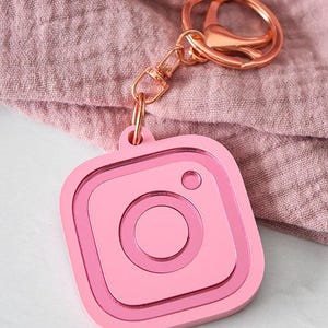 以下が含まれることがあります： ピンクのInstagramロゴのキーホルダーで、ローズゴールドのキーリングが付いています。