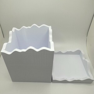 May include: A white, rectangular box and tray set with a wavy, mountain-like edge design. The box is open, revealing its interior. The tray sits beside the box, also with a wavy edge. Both items are made of a thick, white material.