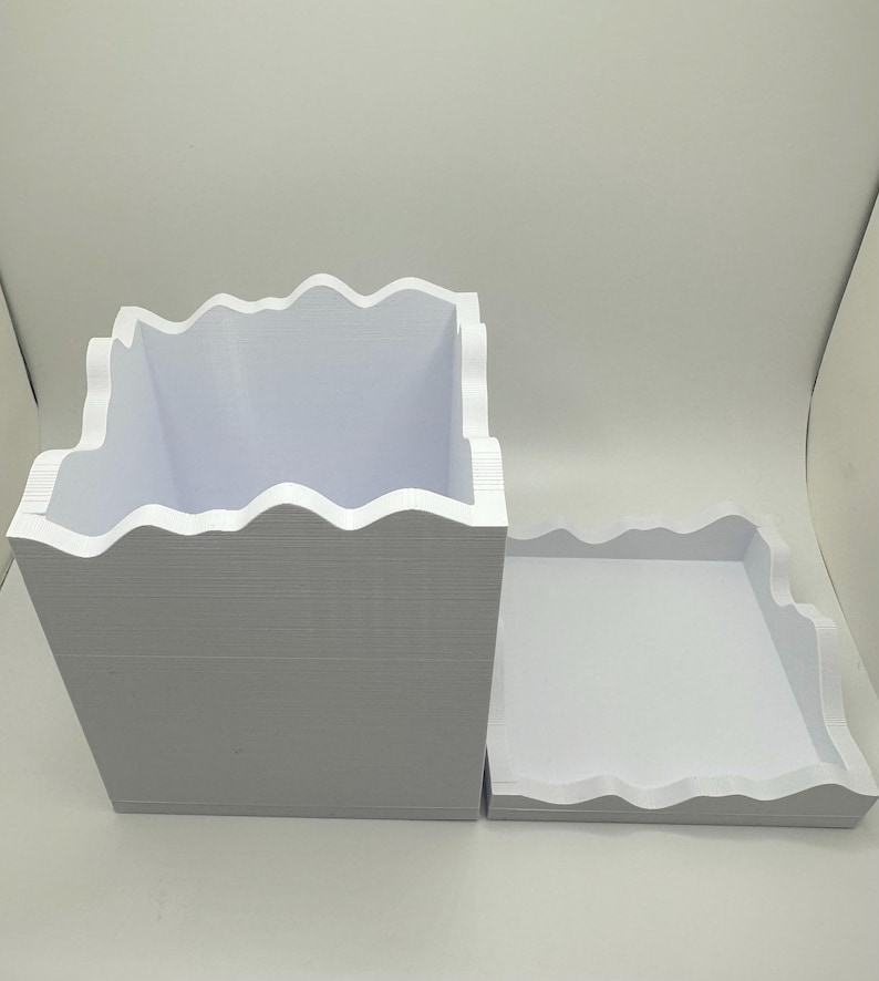 May include: A white, rectangular box and tray set with a wavy, mountain-like edge design. The box is open, revealing its interior. The tray sits beside the box, also with a wavy edge. Both items are made of a thick, white material.