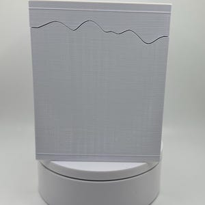 May include: A rectangular, light gray box with a wavy design cut into the top edge. The box has a textured surface and sits on a white, circular base. The background is a plain white.