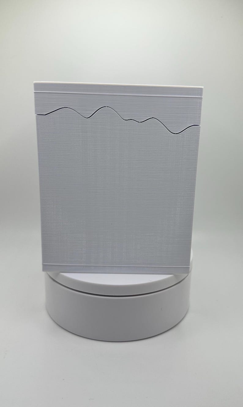 May include: A rectangular, light gray box with a wavy design cut into the top edge. The box has a textured surface and sits on a white, circular base. The background is a plain white.