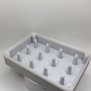 May include: A rectangular gray and white speckled tray featuring a grid of fifteen upright conical pegs. The tray has a smooth, light-colored border and a textured, speckled interior surface. The pegs are arranged in rows and are uniform in shape.