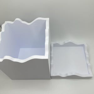 May include: A white, square storage box and a matching tray. Both have a wavy, organic edge design. The box is open, revealing a white interior. The tray sits beside the box, also with a white interior.