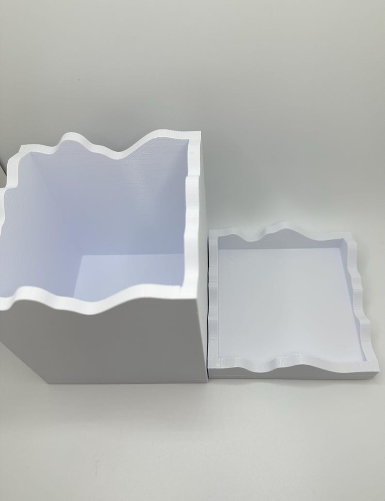 May include: A white, square storage box and a matching tray. Both have a wavy, organic edge design. The box is open, revealing a white interior. The tray sits beside the box, also with a white interior.