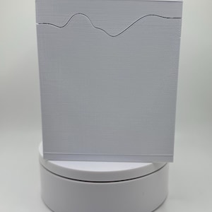 May include: A rectangular, light gray box with a textured surface. A black, wavy line design is etched across the top. The box is displayed on a white, circular stand against a white background.