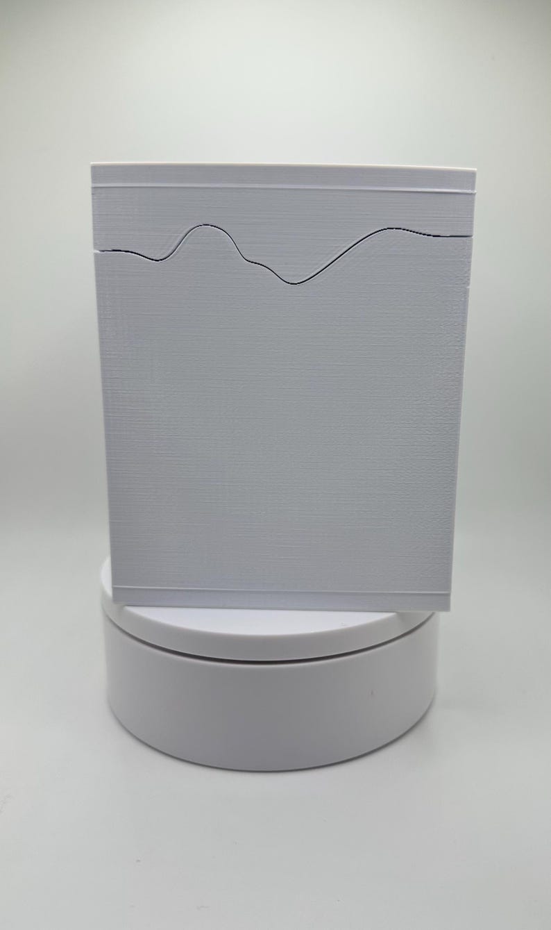 May include: A rectangular, light gray box with a textured surface. A black, wavy line design is etched across the top. The box is displayed on a white, circular stand against a white background.