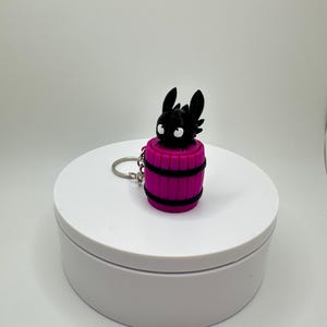 May include: A black dragon figurine with large ears and white eyes sits atop a magenta barrel-shaped keychain. The barrel has black bands around it. The keychain is on a white platform.