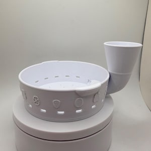 May include: A white, circular, 3D-printed plastic bowl with a perforated base and a side-mounted funnel attachment. The exterior wall features a series of small, embossed shield-shaped icons and rectangular ventilation slots around the perimeter.
