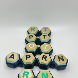May include: A collection of black and gold hexagonal tiles with letters spelling "APRON". The letters are in various colors, including blue, green, and white. The tiles are arranged on a white surface.
