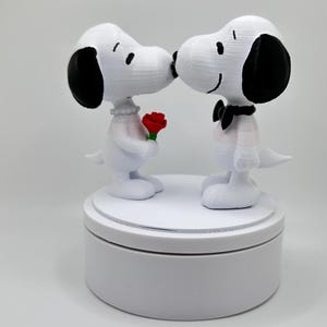 May include: A white figurine set featuring two Snoopy figures in a kissing pose. One Snoopy holds a red rose. The figures have black ears and are on a white circular base. The background is plain white.