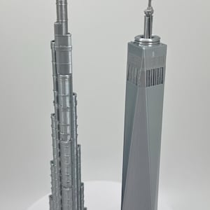 May include: Two gray 3D-printed models of famous skyscrapers. The Burj Khalifa, a tall, slender building, stands next to the One World Trade Center. Both models are detailed and sit on a white surface.