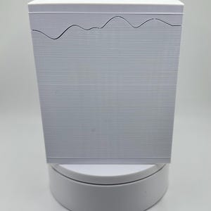 May include: A stack of white paper with a wavy, cut-out design along the top edge. The paper has a textured, horizontal lined pattern. The stack is on a white, circular base, against a plain white background.
