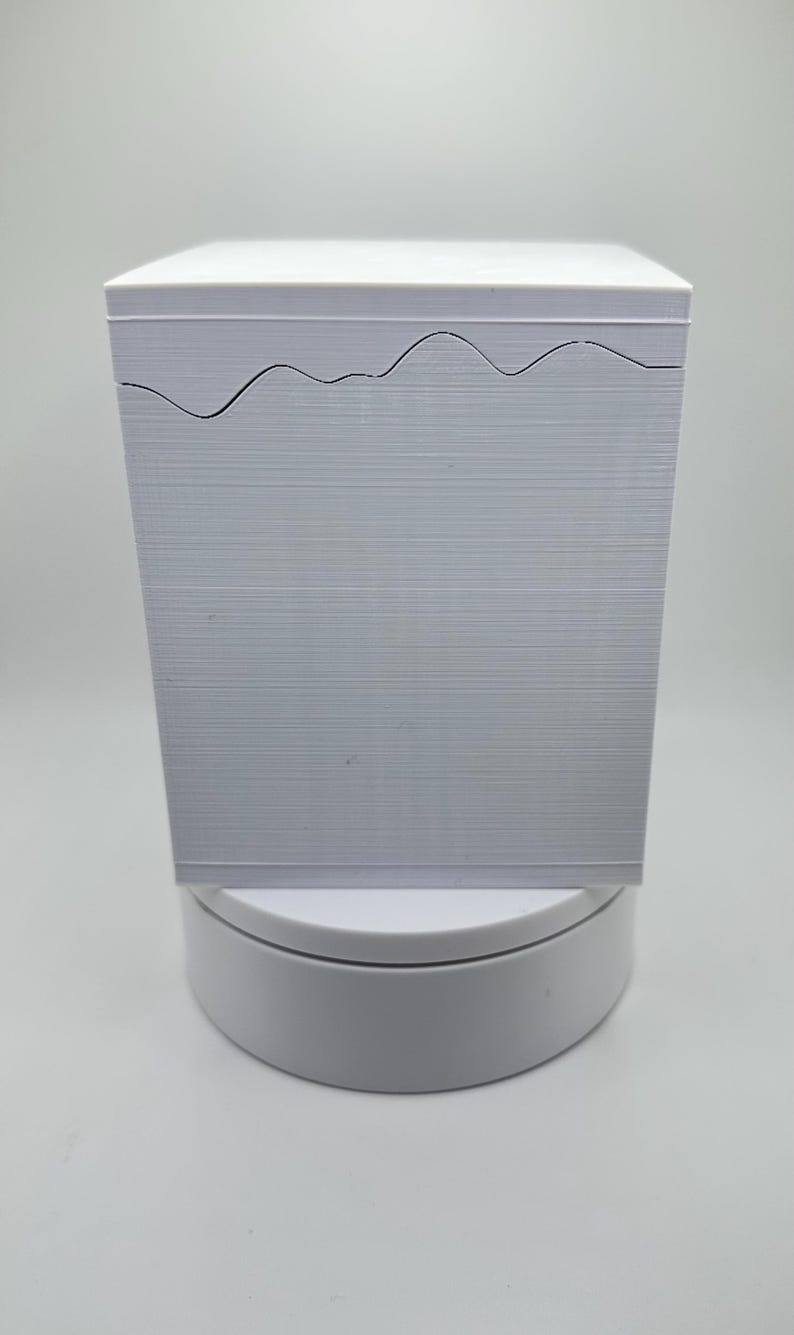 May include: A stack of white paper with a wavy, cut-out design along the top edge. The paper has a textured, horizontal lined pattern. The stack is on a white, circular base, against a plain white background.
