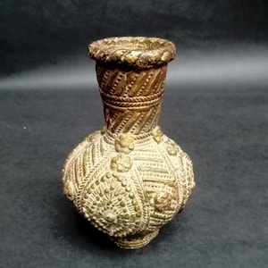 Könnte beinhalten: Verzierte Vase im antiken Stil mit bauchigem Körper und schmalem Hals. Die Vase besteht aus einem metallischen Material mit Gold- und Bronzefinish. Die Oberfläche ist mit komplizierten Mustern und floralen Designs bedeckt.
