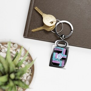 May include: Silver rectangular keychain with a design featuring blue, purple, and black brushstrokes and the text "Going in so many directions with my POTS." Gold keys and a succulent plant are in the background.