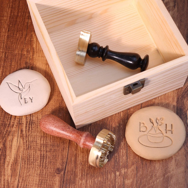 May include: A wooden box with two brass and wooden stamps and two clay impressions. One impression shows a hummingbird with the text "EY" and the other shows a plant with the text "B&H".