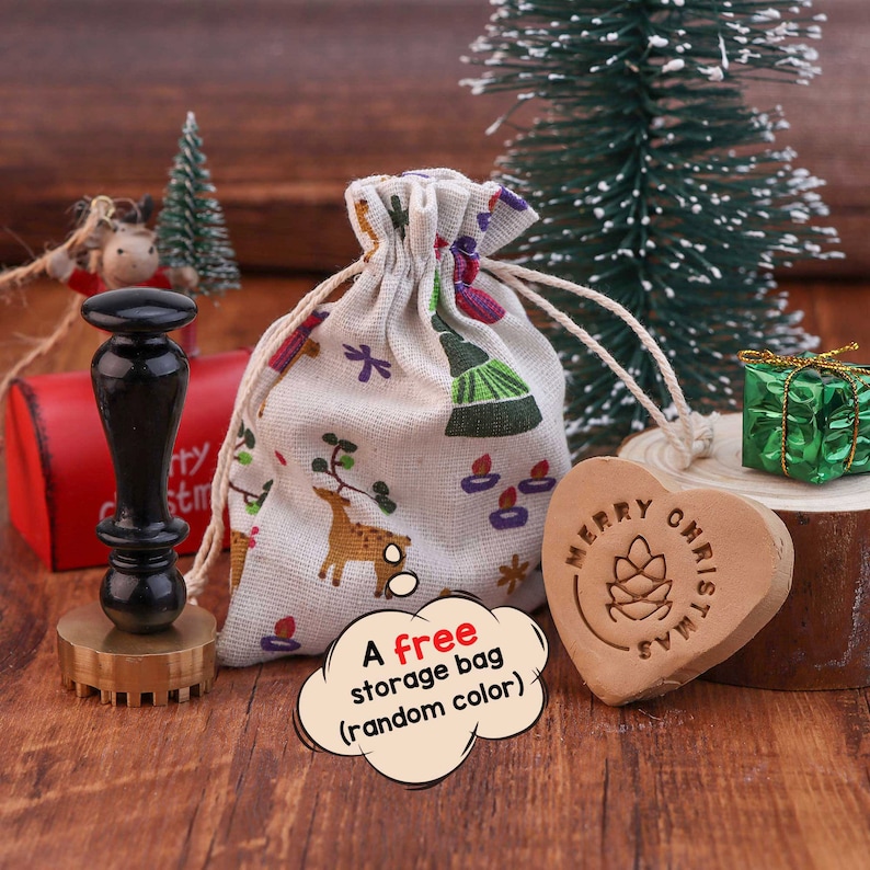 May include: A wooden heart-shaped stamp with the text "Merry Christmas" in a circle around a pine cone design. The stamp is on a wooden surface next to a small white canvas bag with a Christmas design and a small green gift box.