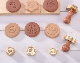 May include: A set of eight round, wooden-handled, brass cookie stamps with various designs. The designs include a ghost, a flower, a dolphin, a leaf, and initials. The stamps are on a white wooden surface.