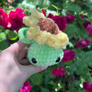 May include: A green crocheted turtle with a yellow crocheted sunflower on its head. The turtle has black eyes and a brown nose. The turtle is being held in a hand against a background of green leaves and pink roses.