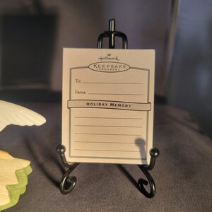May include: A white card with black text that reads "Hallmark Keepsake Ornament" and "Holiday Memory". The card is on a black metal stand.