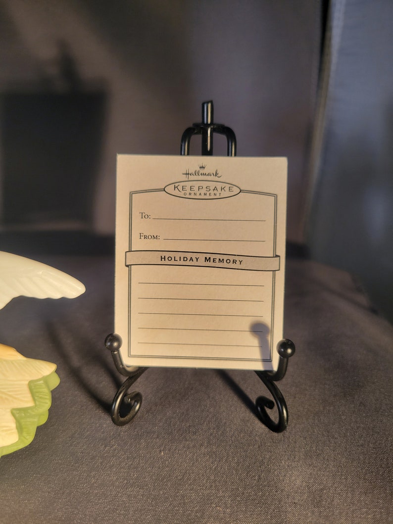 May include: A white card with black text that reads "Hallmark Keepsake Ornament" and "Holiday Memory". The card is on a black metal stand.