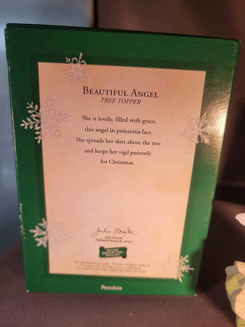 May include: A porcelain angel tree topper with a white dress and poinsettia lace. The angel is holding a white star and has a gold halo. The box features the text "Beautiful Angel Tree Topper" and a poem about the angel. The box is green with snowflakes.
