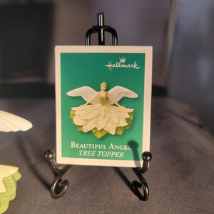 May include: A white angel tree topper with green leaves and a white dress. The angel has wings and is holding a flower. The card reads "Beautiful Angel Tree Topper" and has the Hallmark logo.