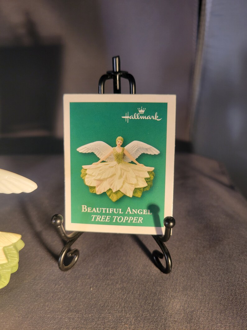 May include: A white angel tree topper with green leaves and a white dress. The angel has wings and is holding a flower. The card reads "Beautiful Angel Tree Topper" and has the Hallmark logo.