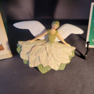 May include: A white porcelain angel figurine with green and white wings and a green and white leaf dress. The angel is wearing a crown of green leaves and has a white face with painted features. The figurine is a tree topper.