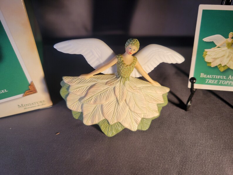 May include: A white porcelain angel figurine with green and white wings and a green and white leaf dress. The angel is wearing a crown of green leaves and has a white face with painted features. The figurine is a tree topper.