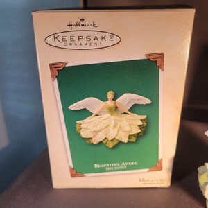 May include: A white and green angel tree topper ornament in a box. The box is white with a green background and the words "Hallmark Keepsake Ornament" and "Beautiful Angel Tree Topper" printed on it.