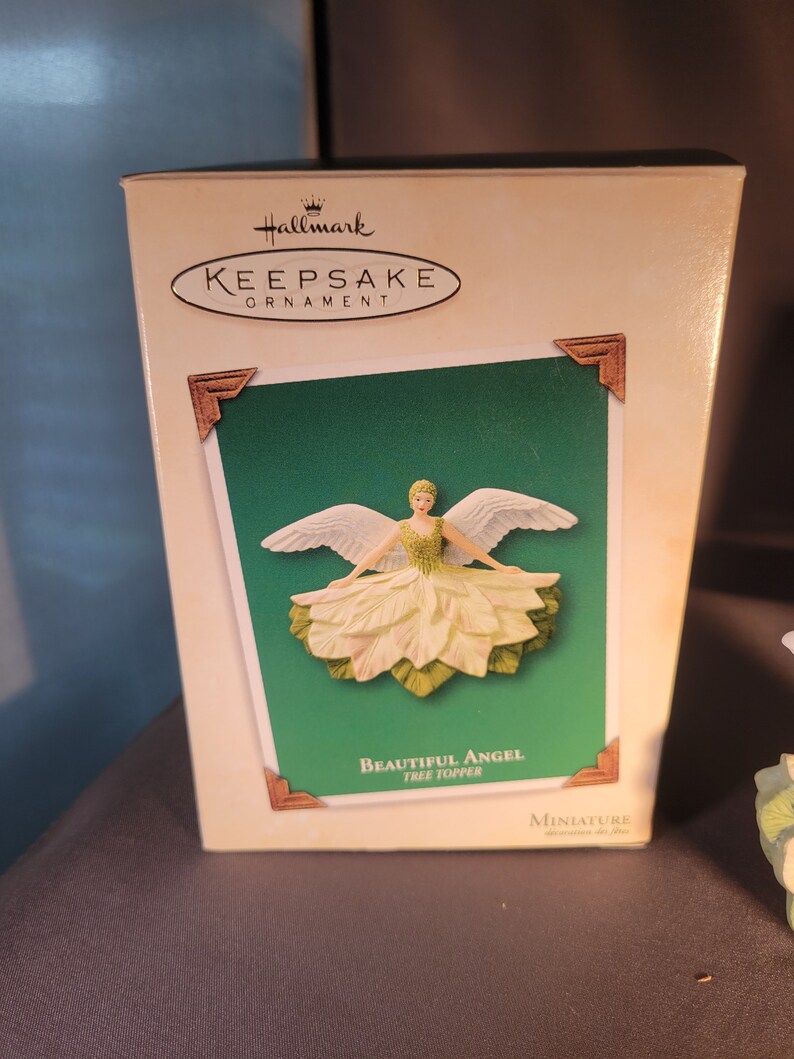 May include: A white and green angel tree topper ornament in a box. The box is white with a green background and the words "Hallmark Keepsake Ornament" and "Beautiful Angel Tree Topper" printed on it.