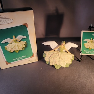 May include: A white angel tree topper with green leaves and wings. The angel is wearing a white dress and has a halo. The ornament is packaged in a Hallmark box with the text "Keepsake Ornament" and "Beautiful Angel Tree Topper".