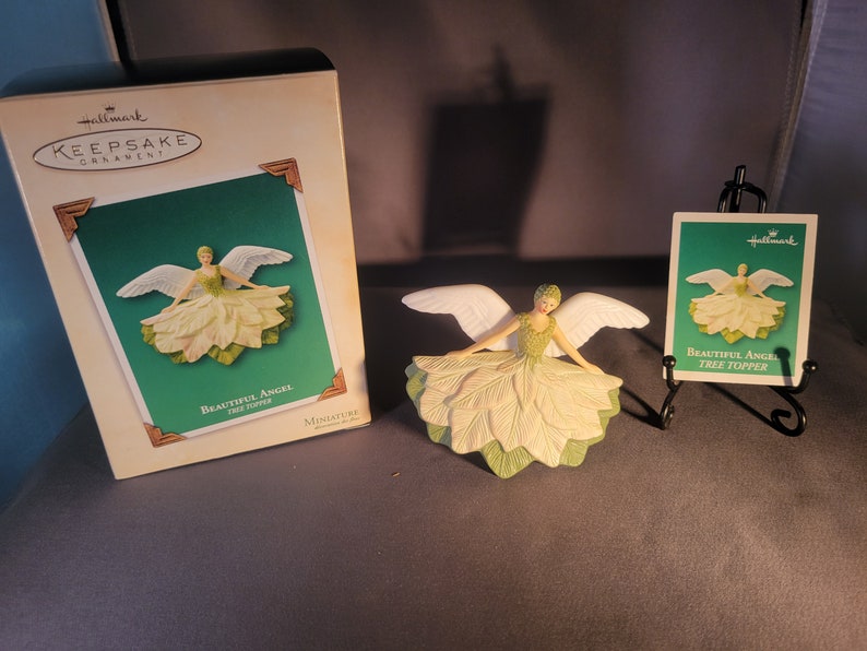 May include: A white angel tree topper with green leaves and wings. The angel is wearing a white dress and has a halo. The ornament is packaged in a Hallmark box with the text "Keepsake Ornament" and "Beautiful Angel Tree Topper".
